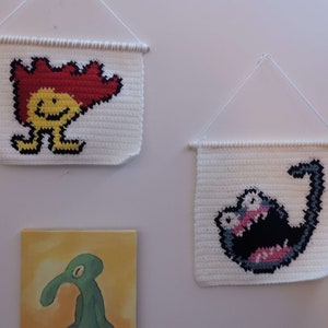 May include: Three wall hangings with crocheted designs. The first is a white crocheted square with a yellow and red cartoon character with a flame-shaped head. The second is a white crocheted square with a black and gray cartoon character with a large mouth. The third is a yellow and orange painting of a green cartoon character with a long nose.