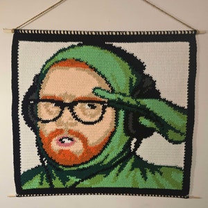 Puede incluir: Un tapiz de pared de ganchillo que representa un retrato de una persona con una sudadera con capucha verde, gafas y barba roja.