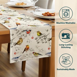 May include: A cream-colored table runner featuring a watercolor design of colorful birds perched on branches with red berries. The runner is placed on a wooden table, with plates and silverware visible. The runner is machine washable, with long-lasting stitching, and sustainably made.