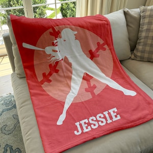 May include: A red fleece blanket with a white silhouette of a softball player holding a bat. The player is standing in a white circle with a pink and white baseball design. The name "JESSIE" is printed in white at the bottom of the blanket.