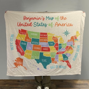 May include: A white fleece blanket with a colorful map of the United States of America. The map includes state names and borders. The blanket has a blue border and the words "Benjamin's Map of the United States of America" at the top.