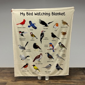 May include: A cream-colored blanket with a colorful illustration of various birds, including a cardinal, bluejay, bald eagle, robin, mourning dove, ruby-throated hummingbird, downy woodpecker, house wren, goldfinch, blackbird, eastern bluebird, northern mockingbird, starling, house sparrow, tufted titmouse, mallard, gambel's quail, red-tailed hawk, pigeon, American robin, Baltimore oriole, and black-capped chickadee. The blanket has the text "My Bird Watching Blanket" at the top.