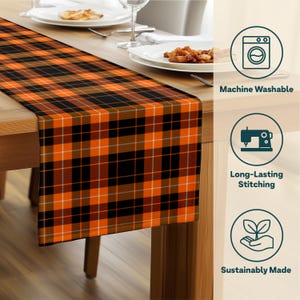 May include: A table runner with a plaid pattern in orange, black, and white. The runner is draped over a wooden table, with plates and silverware visible in the background. The runner is machine washable and sustainably made.