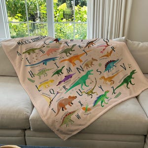 May include: A pink and white blanket with a dinosaur alphabet design. Each letter of the alphabet is paired with a different dinosaur illustration. The blanket features the text "Max's Dinos".