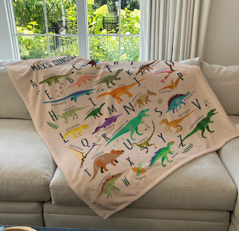 May include: A pink and white blanket with a dinosaur alphabet design. Each letter of the alphabet is paired with a different dinosaur illustration. The blanket features the text "Max's Dinos".