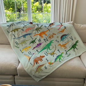 May include: A light green throw blanket with a dinosaur alphabet design. The blanket features colorful illustrations of dinosaurs with their corresponding letters. The text "Max's Dinos" is at the top of the blanket.