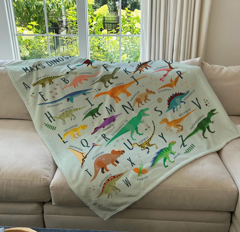 May include: A light green throw blanket with a dinosaur alphabet design. The blanket features colorful illustrations of dinosaurs with their corresponding letters. The text "Max's Dinos" is at the top of the blanket.
