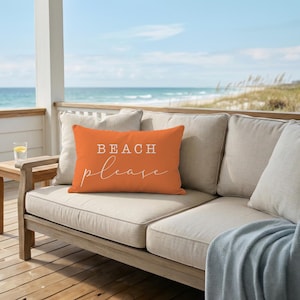 May include: A rectangular orange throw pillow featuring the white text BEACH please in a combination of block and script fonts. The pillow is positioned on a beige outdoor sofa cushion, showcasing a smooth fabric texture and clean edges.