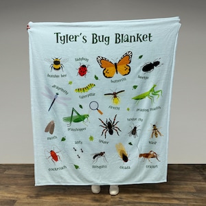 Puede incluir: Una manta azul claro con una variedad de insectos ilustrados en ella. La manta tiene el texto "Tyler's Bug Blanket" en la parte superior. Los insectos incluyen una mariquita, una mariposa, una abeja, una libélula, una oruga, una luciérnaga, una mantis religiosa, una mosca doméstica, un saltamontes, una polilla, hormigas, una araña, una avispa, una cucaracha, un mosquito, una cigarra y un grillo.