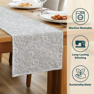 May include: A white table runner with a raised floral pattern, placed on a wooden table. The runner features a detailed design of swirling leaves and vines. The image also shows the text "Machine Washable", "Long-Lasting Stitching", and "Sustainably Made".