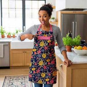 May include: A navy blue apron with a floral pattern in yellow, pink, and blue. The apron has a pink neck strap and a pink label that reads "ALEXANDRA". The apron is being worn in a kitchen setting.