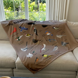 May include: A brown fleece blanket with a colorful illustration of various birds, including a cardinal, hummingbird, bluebird, goldfinch, woodpecker, sparrow, and more. The blanket features the text "My Bird Watch" in a brown font.