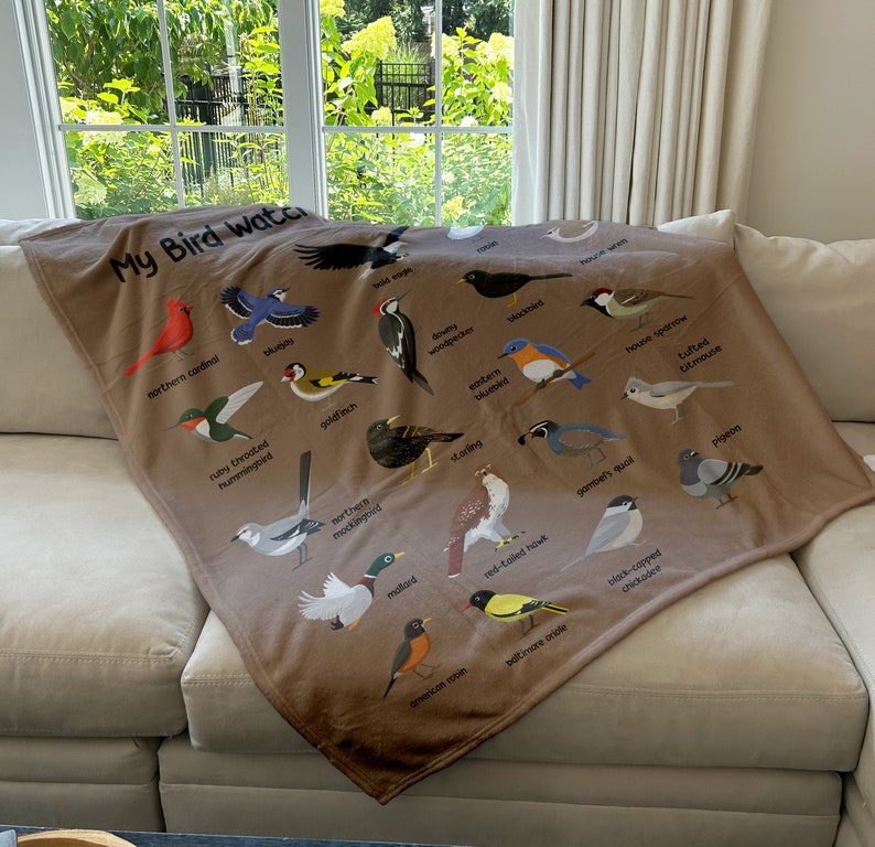 May include: A brown fleece blanket with a colorful illustration of various birds, including a cardinal, hummingbird, bluebird, goldfinch, woodpecker, sparrow, and more. The blanket features the text "My Bird Watch" in a brown font.