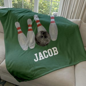 May include: Green blanket featuring a bowling theme. The design includes white bowling pins with red stripes and a black bowling ball. The name "JACOB" is printed in white block letters.