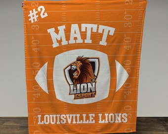 May include: Orange and white fleece blanket with a football design. The football has a lion logo with the text "LION ESPORT". The blanket also has the text "#2 MATT LOUISVILLE LIONS" on it.