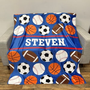 May include: Blue fleece blanket with a sports theme. The blanket features a variety of sports balls, including basketballs, baseballs, footballs, and soccer balls. The blanket also has the name "STEVEN" printed in white letters on a red stripe.