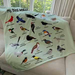 May include: A green fleece blanket with a colorful illustration of various birds. The blanket features the text "My Bird Watch" at the top and names each bird species below its image.