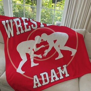 May include: Red fleece blanket with a white wrestling silhouette and the name "ADAM" in white text.