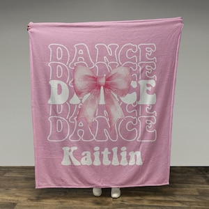 May include: A pink blanket with the word "DANCE" repeated in a wavy font. A pink bow graphic is centered on the blanket. The name "Kaitlin" is printed in white at the bottom. The blanket is held up against a neutral background.
