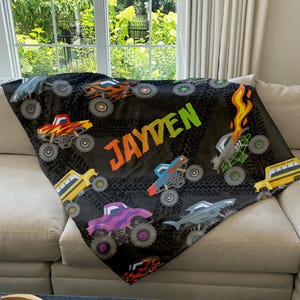 May include: A black fleece blanket with a monster truck theme. The blanket features a variety of colorful monster trucks, tire tracks, and the name "JAYDEN" in a rainbow font.