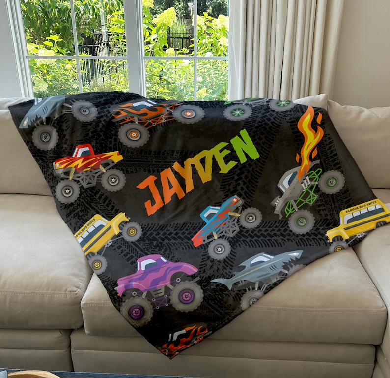 May include: A black fleece blanket with a monster truck theme. The blanket features a variety of colorful monster trucks, tire tracks, and the name "JAYDEN" in a rainbow font.