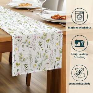 May include: White table runner with a watercolor floral pattern featuring green, yellow, and purple wildflowers. The runner is on a wooden table, with plates and silverware visible. The text "Machine Washable", "Long-Lasting Stitching", and "Sustainably Made" are also visible.