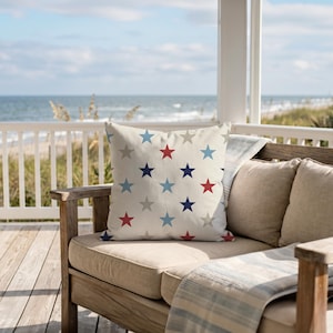 May include: A square decorative pillow with a cream background and a pattern of red, blue, and gray stars. The pillow is placed on a beige cushion of a wooden outdoor sofa, with a view of the ocean in the background.