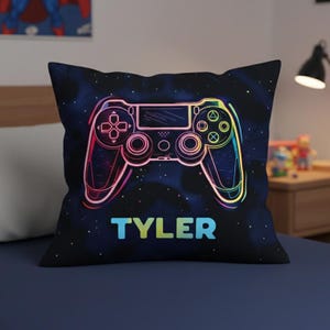 May include: A black square pillow featuring a colorful neon-outlined video game controller design. The name "TYLER" is printed in a gradient of blue, green, and yellow at the bottom of the pillow. The background is a dark blue with small white dots.