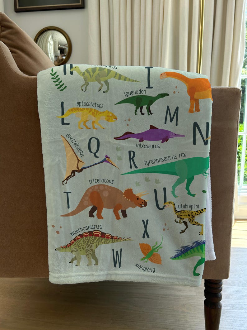 May include: A white blanket with a colorful alphabet featuring dinosaurs. Each letter of the alphabet is paired with a different dinosaur, with the dinosaur's name printed below it. The dinosaurs are illustrated in a cartoon style.