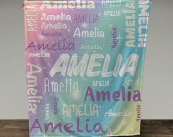 May include: A personalized blanket featuring the name "Amelia" in various fonts and sizes, repeated across a pastel rainbow gradient background. The blanket's colors include shades of pink, purple, blue, and yellow, creating a soft, dreamy aesthetic. The blanket is held up by a person.