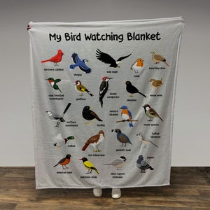 May include: A white fleece blanket with a colorful illustration of different bird species. The blanket features the text "My Bird Watching Blanket" at the top. Each bird is labeled with its name.