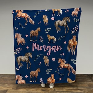 May include: A navy blue fleece blanket with a repeating pattern of brown horses and pink flowers. The blanket has the name "Morgan" written in pink letters.