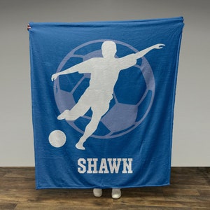 May include: A blue fleece blanket with a white silhouette of a soccer player kicking a ball. The player is inside a white soccer ball with a blue outline. The name "SHAWN" is printed in white letters below the soccer ball.