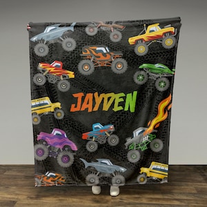May include: A black fleece blanket with a monster truck theme. The blanket features a variety of colorful monster trucks, including a school bus, a purple truck, and a blue truck. The blanket also has the name "JAYDEN" printed on it.