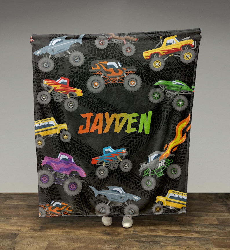 May include: A black fleece blanket with a monster truck theme. The blanket features a variety of colorful monster trucks, including a school bus, a purple truck, and a blue truck. The blanket also has the name "JAYDEN" printed on it.