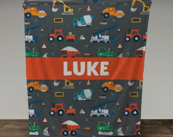 Puede incluir: Una manta de franela gris con un patrón de vehículos de construcción. La manta tiene una franja roja con el nombre "LUKE" en letras blancas.