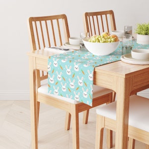 May include: A light blue table runner with a repeating pattern of white bunnies and orange carrots. The runner is on a wooden table with two chairs.