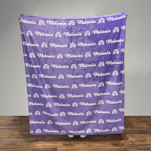 May include: Purple fleece blanket with a white repeating pattern of the name "Melanie" with a rainbow arc above each name.