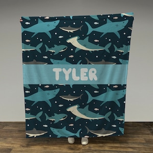 May include: A blue and white personalized fleece blanket with a shark and hammerhead shark pattern. The name "TYLER" is printed in white on a blue banner.