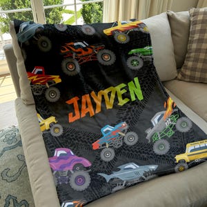 May include: A black fleece blanket with a monster truck theme. The blanket features a variety of colorful monster trucks, including a school bus, and the name "JAYDEN" in a rainbow font.