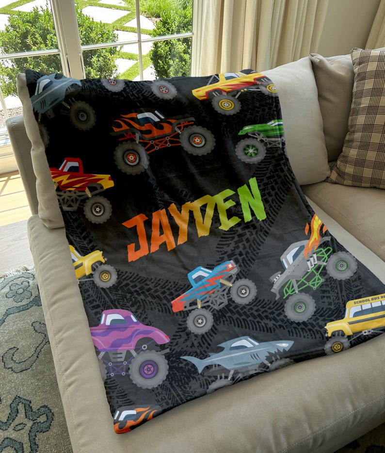 May include: A black fleece blanket with a monster truck theme. The blanket features a variety of colorful monster trucks, including a school bus, and the name "JAYDEN" in a rainbow font.