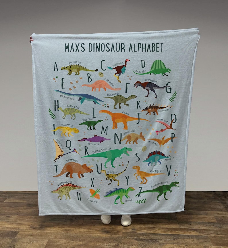 May include: A light blue blanket with a dinosaur alphabet printed on it. Each letter of the alphabet is paired with a different dinosaur, with the dinosaur's name printed below it. The blanket has the words "Max's Dinosaur Alphabet" printed at the top.