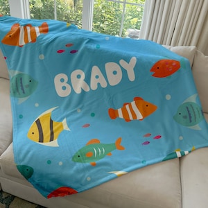May include: A light blue blanket featuring colorful cartoon fish and the name "BRADY" in large white letters. The blanket has a soft texture and is decorated with various fish designs in orange, yellow, green, and blue.