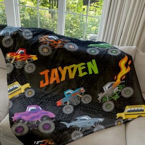 May include: A black fleece blanket with a monster truck theme. The blanket features a variety of colorful monster trucks, tire tracks, and the name "JAYDEN" in green letters.