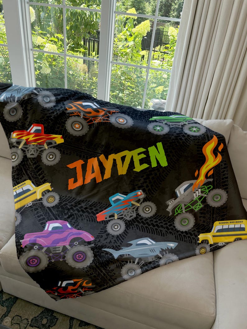 May include: A black fleece blanket with a monster truck theme. The blanket features a variety of colorful monster trucks, tire tracks, and the name "JAYDEN" in green letters.