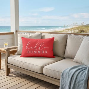 May include: A rectangular throw pillow in a vibrant coral red color featuring the white text "hello SUMMER" printed on the front. The word "hello" is written in a cursive script font, while the word "SUMMER" is in a bold, block capital font.