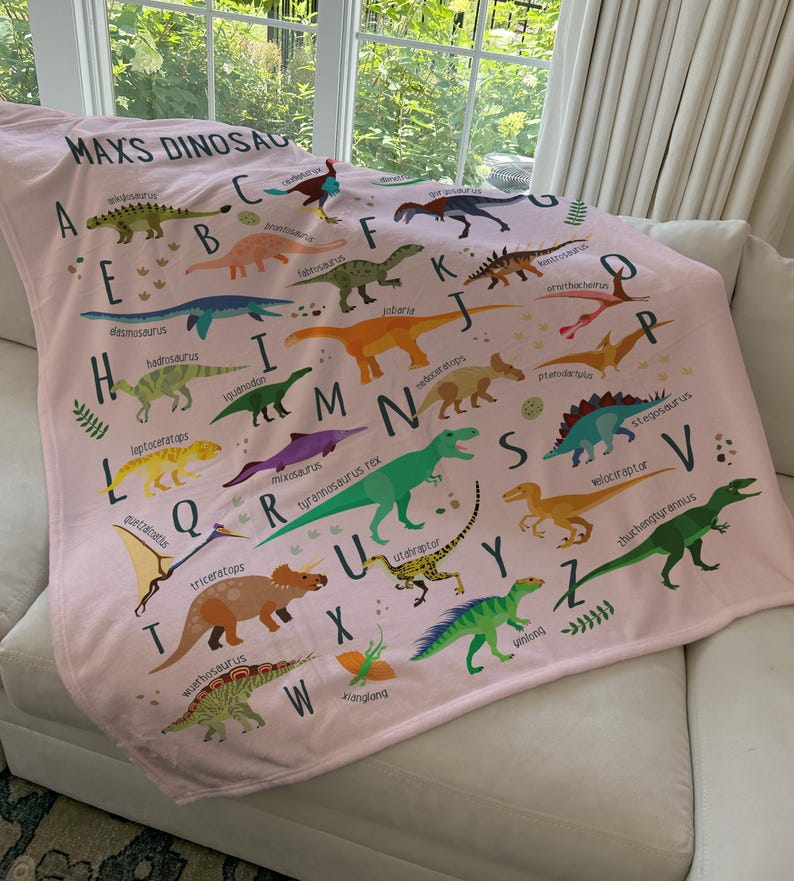 May include: A pink fleece blanket with a dinosaur alphabet design. Each letter of the alphabet is paired with a different dinosaur and its name. The blanket features a variety of dinosaurs, including a stegosaurus, a velociraptor, a triceratops, and a tyrannosaurus rex.