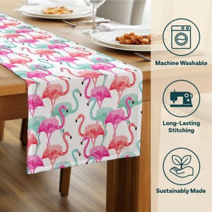 May include: A white table runner featuring a repeating pattern of pink and teal flamingos. The flamingos are in various poses against a white background. The runner is on a wooden table, with plates and silverware visible.