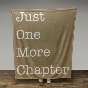 May include: A tan fleece blanket with the text "Just One More Chapter" printed in white.