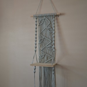 May include: A grey macrame wall hanging with a wooden shelf. The macrame has a geometric pattern and is finished with tassels.